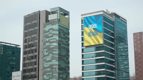 LED-Screen-On-High-rise-Building-Showing-PEACE-NO-WAR-Over-Ukrainian-Flag-During-Russo-Ukrainian-War-In-Seoul,-South-Korea