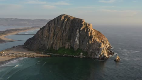 aerial-drone-circling-Morro-Bay-Rock-Beach-in-California-USA-during-sunset-as-pacific-ocean-waves-crash-onto-the-sandy-beach-on-a-summer-afternoon