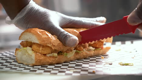 Chef-uses-red-serrated-knife-to-cut-fried-shrimp-poboy-sandwich-in-half,-slow-motion-close-up-4K