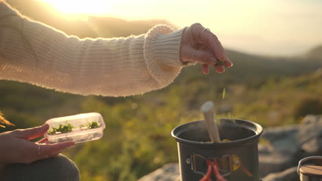 Close-up-shot-revealing-a-woman-cooking-food-in-a-black-pot-on-top-of-a-mountain-at-sunset