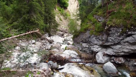 Aerial-dolly-flying-downstream-a-creek-flowing-through-a-mountain-forest,-descending-view