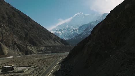 Drone-Rising-Up-Over-Road-Past-Shadow-Of-Valley-To-Reveal-Snow-Capped-Mountains-Of-Hunza-Valley