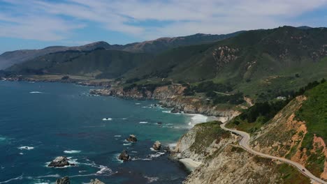 Wide-aerial-view-of-a-winding-curvy-highway-road-along-Route-1-on-the-coast-of-Big-Sur-California-with-large-mountain-cliffs-and-waves-crashing-into-the-rocks-below