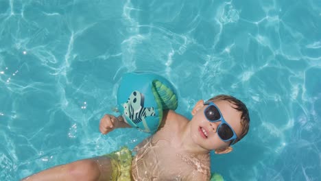Cooler-Junge,-Der-In-Die-Kamera-Blickt,-Sonnenbrille-Und-Schwimmkörper-Trägt,-Unberührtes-Wasser-Im-Pool,-Von-Oben-Nach-Unten