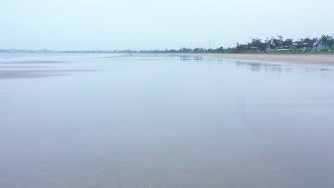 traveling-ground-shot-of-watery-calm-shore,-drone-rises-to-showcase-the-beach,-mellow-gloomy-sky,-single-person-on-beach,-trees-in-distance,-calm-tranquil-vibe,-empty-beach,-cloudy-day-sky,-peaceful