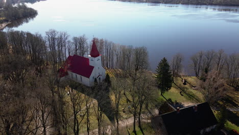 Aerial-view-of-Lielvarde-Lutheran-church-at-the-bank-of-Daugava-river,-white-church-with-red-roof,-leafless-trees,-sunny-spring-day,-wide-shot-moving-forward-camera-tilt-down