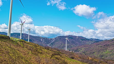 Static-view-of-white-wind-turbines-moving-in-timelapse-over-a-hilly-terrain-with-white-clouds-passing-by-on-a-bright-sunny-day