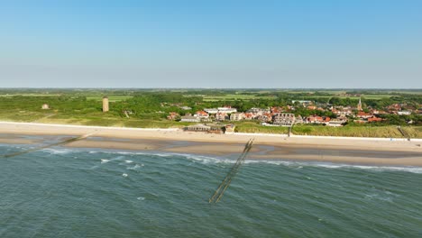 Long-aerial-shot-flying-over-the-sea-and-beach-towards-a-picturesque-little-rural-coastal-town-with-orange-rooftops-on-a-cloudless-summer-day
