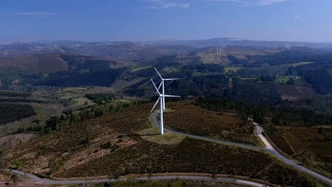 Spinning-wind-turbines-lined-up-on-a-hill-with-forests-connected-by-roads,-plus-wind-turbines-in-the-background-on-the-horizon
