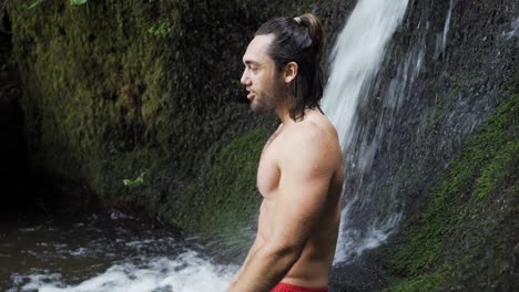 Young-topless-fit-man-taking-a-bath-in-a-spring-and-enjoing-the-fresh-water-from-a-beautiful-Waterfall