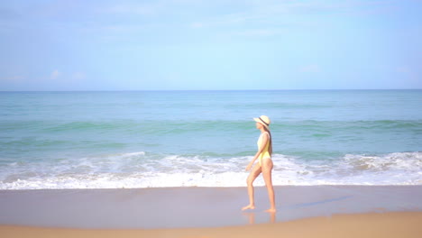 Adult-asian-woman-in-yellow-swimwear-walks-by-the-sea-on-a-sandy-beach