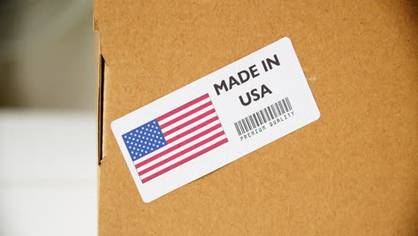 Manos-Aplicando-Etiqueta-De-Bandera-Hecha-En-EE.UU.-En-Una-Caja-De-Cartón-De-Envío-Con-Productos