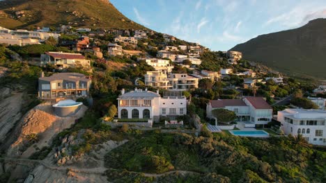 Expensive-white-mansions-on-mountain-cliff-at-Llandudno-Beach-Cape-Town-South-Africa-at-sunset,-aerial
