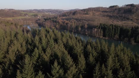Drone-flight-over-a-pine-forest-that-surrounds-a-blue-lake-in-the-German-countryside-at-sunset