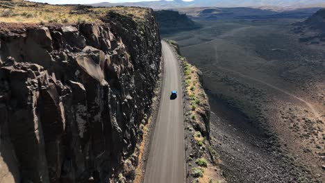 Wide-drone-shot-of-a-bright-blue-car-driving-on-a-rocky-highway