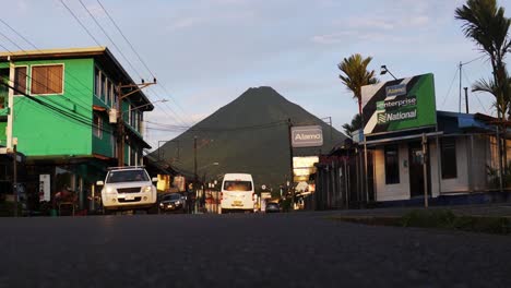 Cars-passing-by-at-a-street-in-la-fortuna,-costa-rica-with-the-volcano-el-arenal-in-the-background