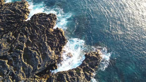 Top-down-drone-footage-of-a-cooled-lava-beach-with-blue-tropical-water-and-waves-during-sunset