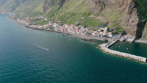 Seaside-town-below-coastal-cliffs,-boat-cruising-to-harbor,-Madeira