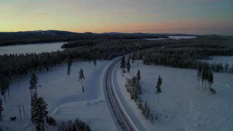 Aerial-view-over-long-curving-snowy-frozen-road-between-Lapland-winter-forest-landscape-at-sunrise