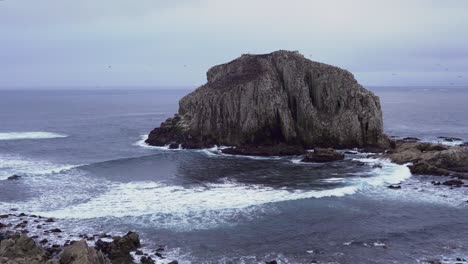 Foamy-sea-waves-and-flock-of-birds-resting-in-Peña-Blanca-rocky-islet-on-a-cloudy-day,-Algarrobo,-Chile