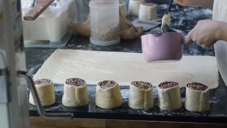 Melted-butter-brushed-onto-freshly-rolled-sheet-of-dough-on-kitchen-table-top-with-cinnamon-rolls-on-display-nearby