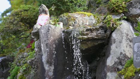 Estatua-De-Jizo-Cubierta-De-Musgo-Y-Agua-Fluyendo-Sobre-Rocas-En-Un-Jardín-Japonés-En-Cámara-Lenta