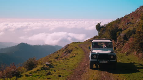 Tracking-shot-of-white-jeep-driving-on-dangerous-rural-road-on-high-mountain-over-clouds