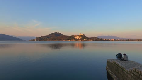 Romantic-couple-sit-on-jetty-edge-over-Maggiore-lake-and-look-at-Angera-castle-in-background-at-sunset,-Italy