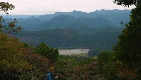 Hiker-walking-past-landscape-of-forested-mountains-and-distant-Torii-gate,-Kumano-Kodo