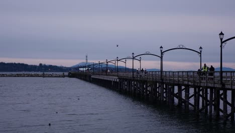 Pan-with-people-walking-on-a-long-pier-leading-towards-a-break-water-in-ocean-during-the-evening-clouds-at-dusk