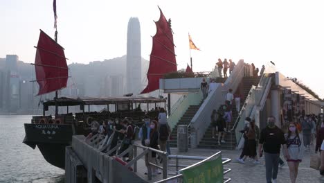 Victoria-Harbour-waterfront-as-a-wooden-red-sailed-junk-boat-based-on-ancient-Chinese-sailing-ships,-now-used-as-a-touristic-attraction,-is-seen-in-front-of-the-Hong-Kong-Island-skyline