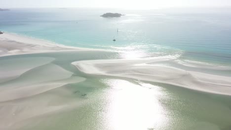 Rotating-sun-beam-aerial-over-shallow-sand-lagoon,-sailboats-beyond