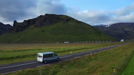 Furgoneta-Conduciendo-Por-Una-Carretera-Asfaltada-Pasando-Por-Campos-Verdes-Con-Vistas-A-La-Montaña-En-Islandia