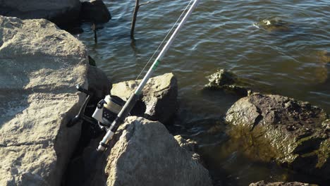 Fishing-rod-set-up-waiting-for-fish-to-bite-bait,-rocky-lake-coastline