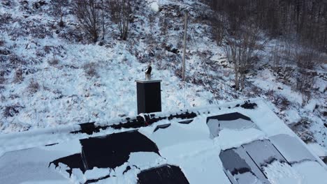 Smoke-coming-from-wood-stove-chimney-while-moving-bacwards-to-reveal-snow-covered-solar-roof-and-frozen-landscape---Norway-winter-season-aerial