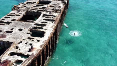 Drone-Aerial-View,-Female-Person-Jumping-From-Shipwreck-in-Turquoise-Sea-Water