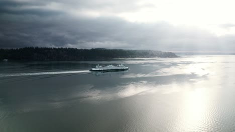Commuter-ferry-cruises-along-dark-water-reflecting-streaming-sunlight-and-gloomy-clouds,-aerial
