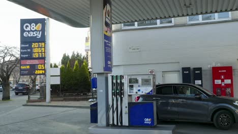 Teures-Auto,-Das-Die-Q8-Tankstelle-Verlässt,-Vor-Einer-Interaktiven-Tafel,-Auf-Der-Die-Höchsten-Kraftstoffpreise-Aller-Zeiten-Angezeigt-Werden