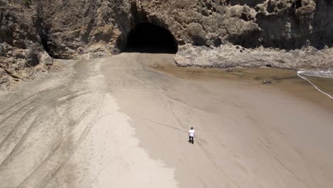 Toma-Aérea-De-Una-Persona-Caminando-Hacia-La-Cueva-Cerca-De-La-Playa-De-Bethells-En-Nueva-Zelanda