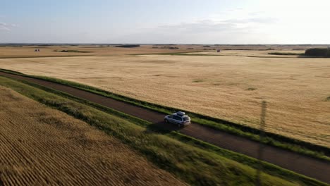 Aerial-4k-shot-of-silver-car-driving-along-dusty-country-road-during-sunset-in-the-prairie