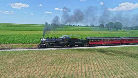 An-Aerial-View-of-a-Steam-Engine-Puffing-Smoke-and-Steam-with-Passenger-Coaches-Traveling-on-a-Single-Track-Thru-Planted-Fields-and-Farmland-Countryside-on-a-Beautiful-Cloudless-Spring-Day