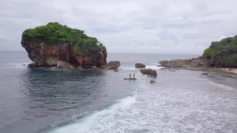 Aerial-view-of-two-people-standing-on-rock-in-bay-of-Jungwok-Beach-in-Indonesia-during-cloudy-day---Giant-Boulder-with-growing-trees-in-background