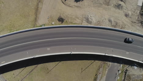 Dark-grey-silver-car-driving-over-bridge-from-left-to-right-of-frame-from-a-static-aerial-perspective