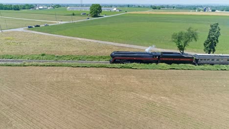 An-Aerial-View-of-a-Steam-Engine-Puffing-Smoke-and-Steam-with-Passenger-Coaches-Traveling-on-a-Single-Track-Thru-Trees-and-Farmland-Countryside-on-a-Beautiful-Cloudless-Spring-Day