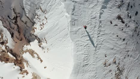 overhead-drone-shot-of-hiker-walking-along-a-snowy-mountain-ridge-overlooking-a-steep-drop