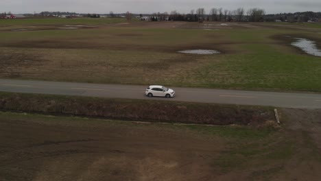 white-car-2019-toyota-corolla-hatchback-driving-along-country-farm-road-surrounded-by-grass-brown-fields-mountains-in-farmland-Abbotsford-BC-Aerial-wide-leading-tracking-car-accelerates-side-profile