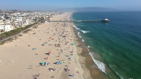 Mittagsseitenansicht-Des-Strandes-Von-Manhattan-Voller-Strandgänger,-Die-In-Kalifornien,-USA,-Schwimmen-Und-Surfen-–-Nach-Vorne-Schwenkende-Luftaufnahme