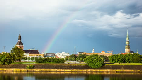 Zeitrafferaufnahme-Eines-Wunderschönen-Regenbogens-über-Der-Stadt-Riga-An-Bewölkten-Tagen,-Lettland---Wunderschöne-Kirchen,-Fluss-Daugava-Und-Fahrende-Autos-Auf-Der-Straße