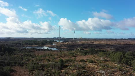 Wind-turbines-at-Lindesnes-wind-farm-Norway---Beautiful-approaching-aerial-over-treetops-with-two-wind-turbines-in-center-and-sky-reflections-in-lake---One-turbine-running-and-one-stopped