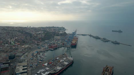 Aerial-dolly-in-of-Valparaiso-hillside-city-and-Sea-Port,-boats-docked-and-cargo-ship-near-cranes-at-sunset,-Chile